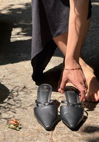 Schwarze spitze Mules aus glattem Material mit zwei dünnen Riemen über dem Fußrücken. Die Oberfläche befindet sich auf einem strukturierten Steingrund.