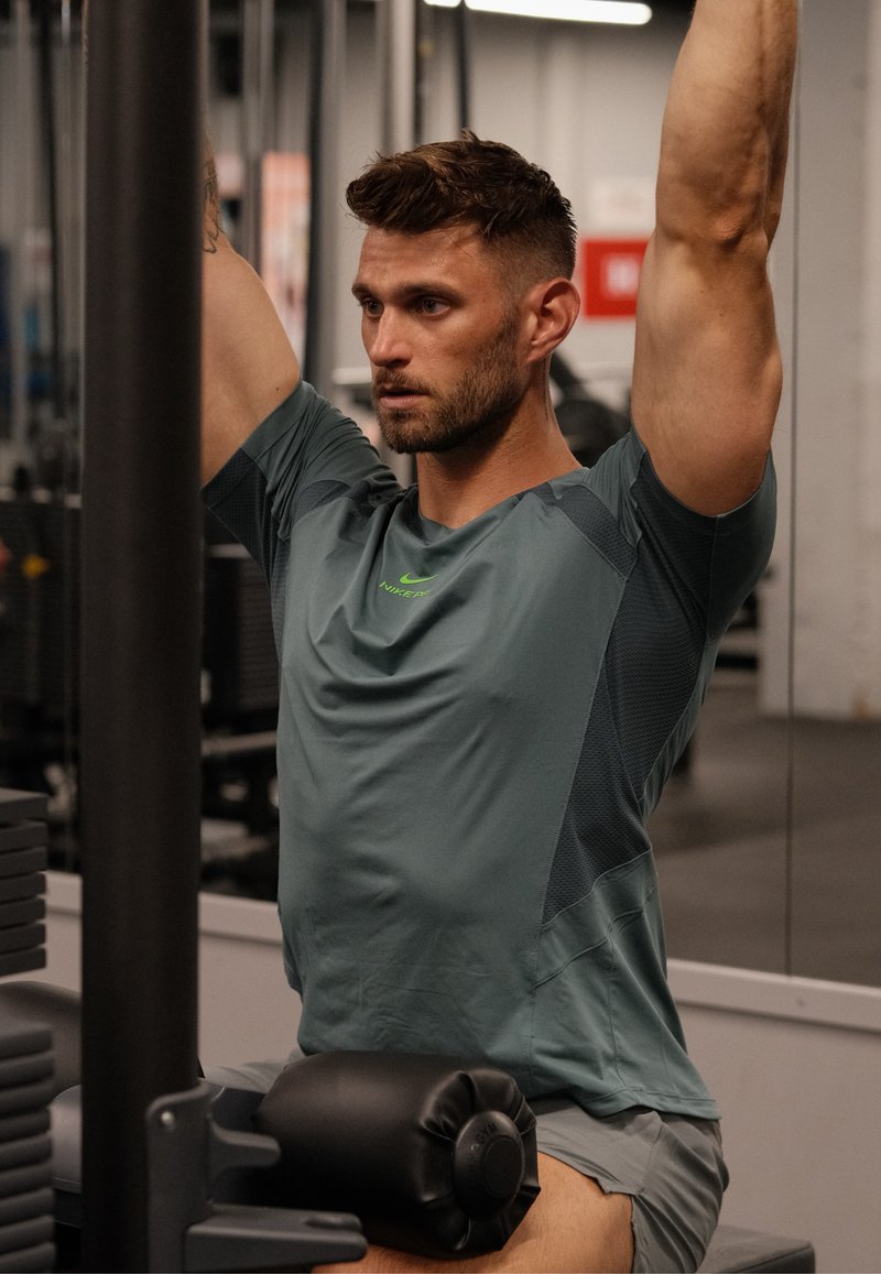 Homme portant un t-shirt gris Nike s'exerce en tirant vers le bas une barre sur un appareil de gym, assis avec une expression concentrée.