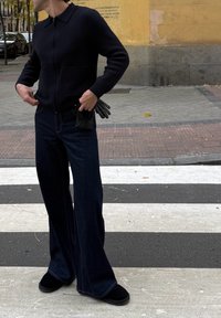 Personne portant un pull noir à fermeture éclair et un jean large foncé tenant des gants noirs, debout sur un passage piéton en ville près d'un mur jaune.