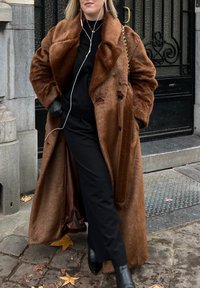 Γυναίκα με μακρύ καφέ παλτό από γούνα, μαύρο παντελόνι και μπότες, στέκεται σε πεζοδρόμιο καλυμμένο με φύλλα κοντά σε μια μαύρη μεταλλική πόρτα, φορώντας ακουστικά.