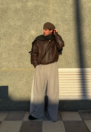 Chaqueta de cuero marrón, pantalones de chándal grises holgados con rayas blancas, bufanda negra y boina marrón. De pie contra una pared gris con textura.