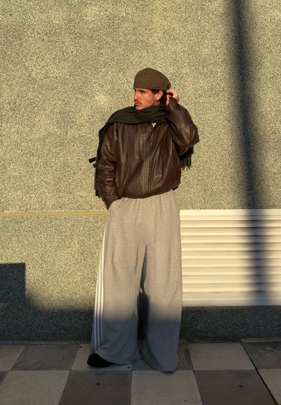 Chaqueta de cuero marrón, pantalones de chándal grises holgados con rayas blancas, bufanda negra y boina marrón. De pie contra una pared gris con textura.