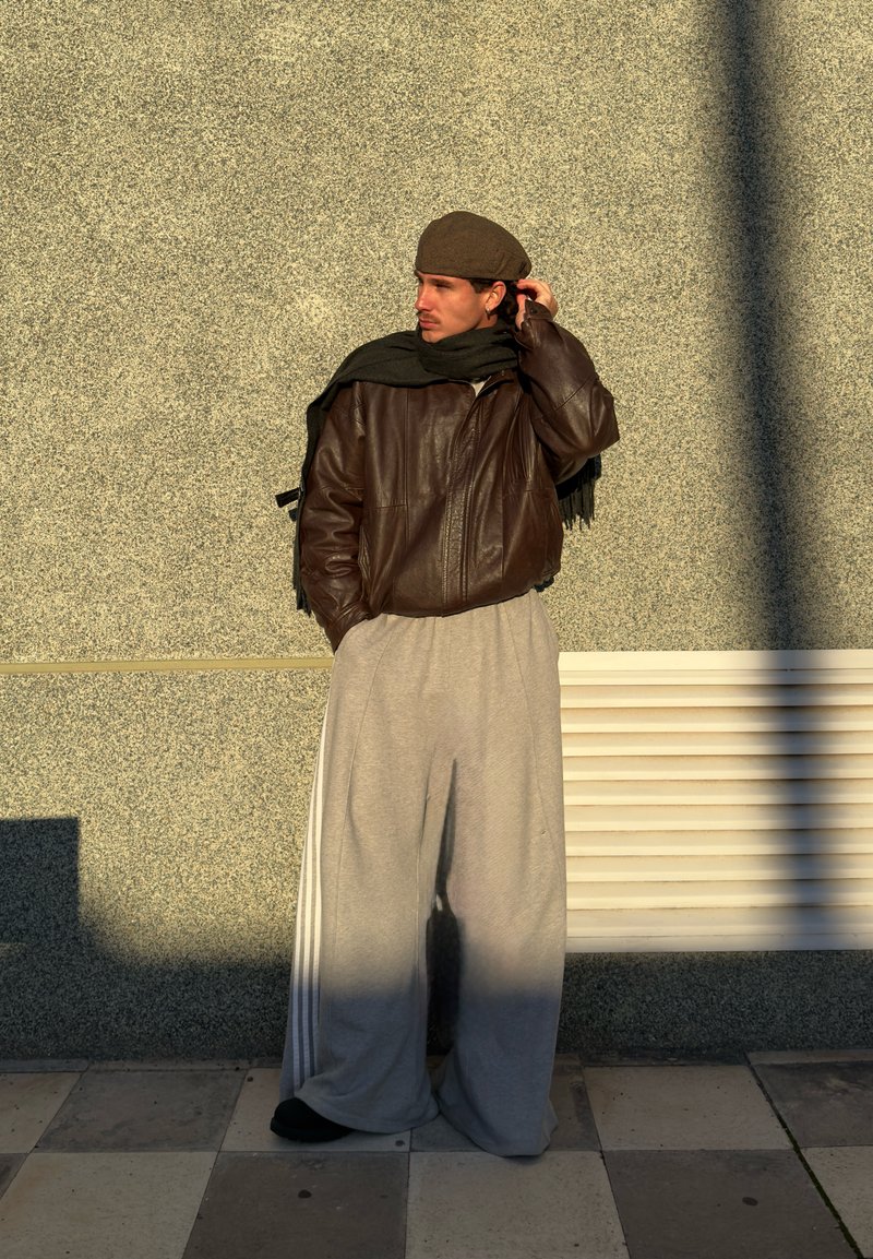Veste en cuir marron, pantalon de survêtement ample gris avec des bandes blanches, écharpe noire et béret marron. Debout contre un mur gris texturé.