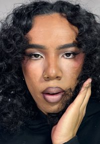 Portrett med krøllete svart hår, sølvsminkeaksenter og en gyllen ring i nesen. Personen hviler en hånd mot ansiktet, og viser frem en matt leppefarge.