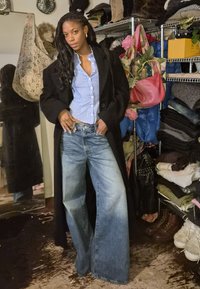 Mannequin portant un pardessus noir, une chemise à rayures bleu clair à boutons, et un jean large délavé, debout devant un arrière-plan encombré.
