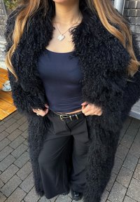 Femme portant un manteau en fourrure noire texturé, un débardeur bleu marine, un pantalon large noir, une ceinture noire et un collier papillon, debout en plein air.