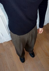 Maglia blu navy a maniche lunghe abbinata a pantaloni larghi marroni a righe sottili e scarpe nere. Il pavimento è in legno chiaro, con uno sfondo bianco visibile.