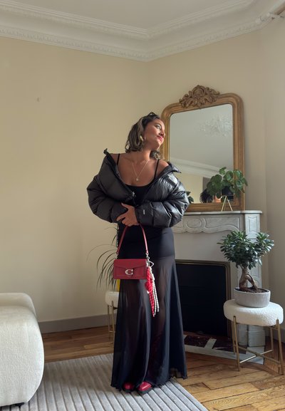 Una mujer vestida con un vestido negro y una chaqueta acolchada sostiene un bolso rojo, de pie cerca de taburetes blancos, plantas y un espejo decorativo con marco dorado en la sala de estar.