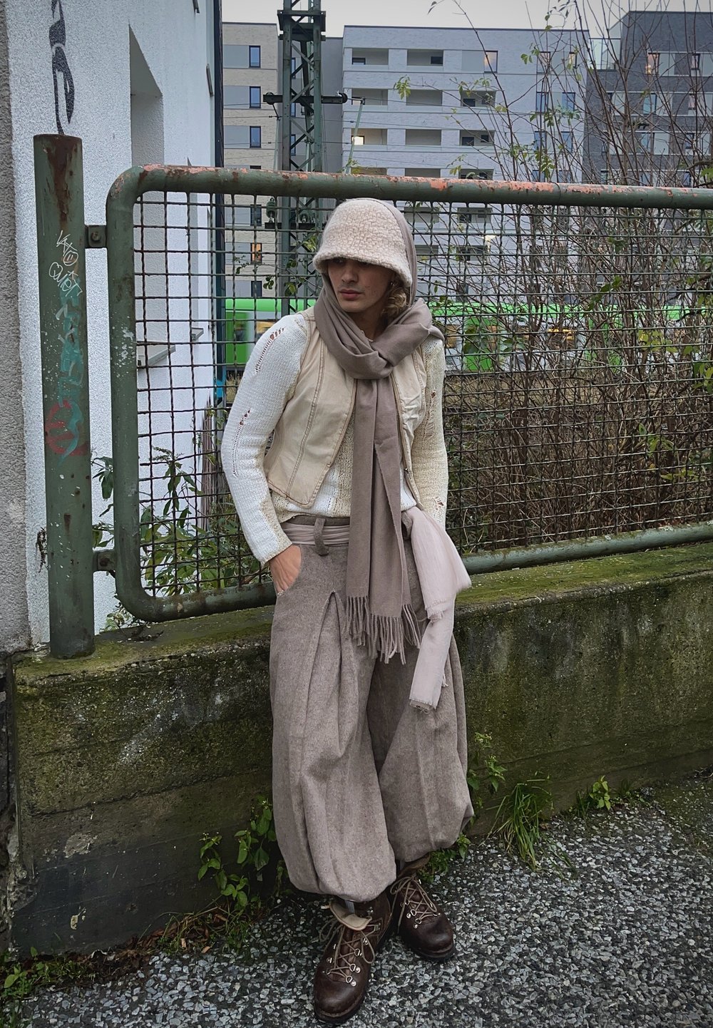 Μπεζ κοντό γιλέκο πάνω από έναν λευκό πλεκτό πουλόβερ, σε συνδυασμό με φαρδιά γκρι παντελόνια και καφέ μαντήλι. Καφέ μποτάκια με κορδόνια. Υφασμάτινο φόντο.