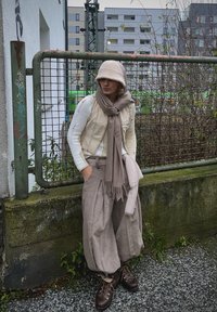 Μπεζ κοντό γιλέκο πάνω από έναν λευκό πλεκτό πουλόβερ, σε συνδυασμό με φαρδιά γκρι παντελόνια και καφέ μαντήλι. Καφέ μποτάκια με κορδόνια. Υφασμάτινο φόντο.