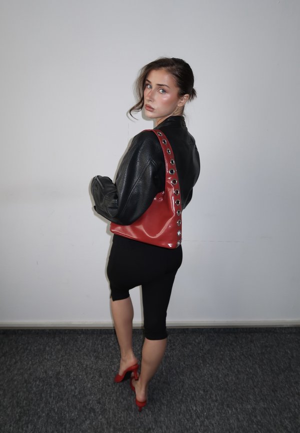 Woman in black leather jacket and skirt wearing red heels, carrying red shoulder bag with metal eyelets, standing against plain white wall.
