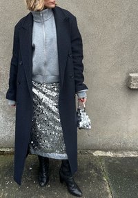 Long manteau bleu marine sur un sweat-shirt gris, jupe en sequins argentés et bottes en cuir noir. Petit sac à main à motifs complète la tenue.