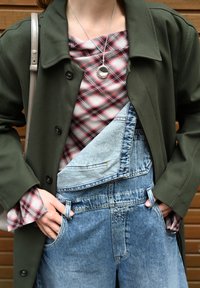 Donkergroene jas over een geruite rood-witte top, gecombineerd met lichtblauwe denim overall met zijzakken en een relaxed fit. Zilveren ketting zichtbaar.
