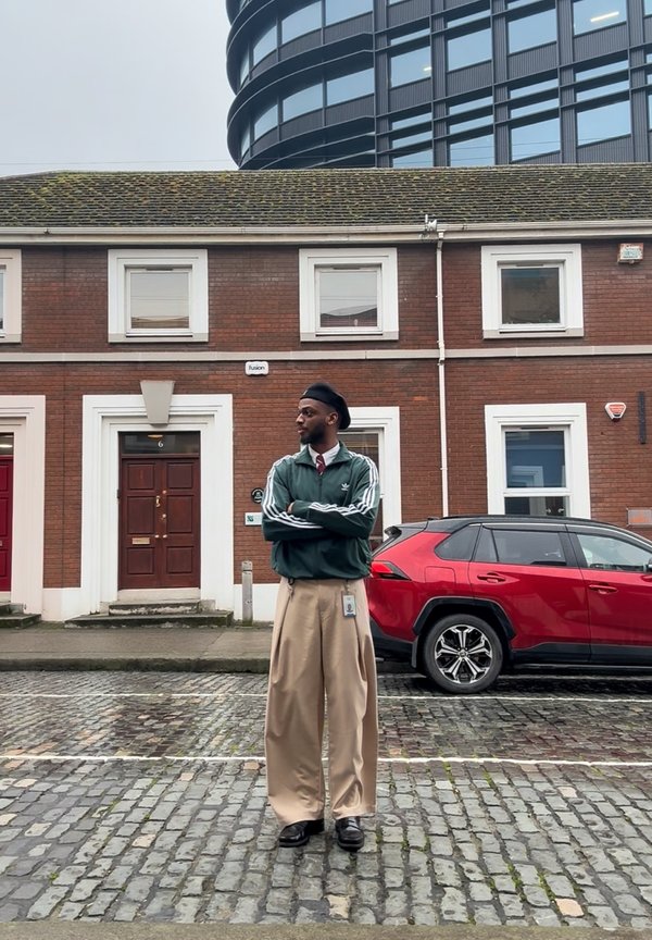 Homme en veste verte, pantalon large beige et béret noir debout dans une rue pavée, avec une voiture rouge et un immeuble en brique en arrière-plan.