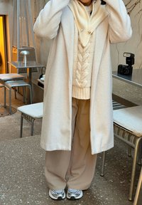 Manteau en laine beige porté sur un pull crème à torsades, assorti à un pantalon beige à jambes larges et des baskets blanches. Textures lisses, coupe décontractée.