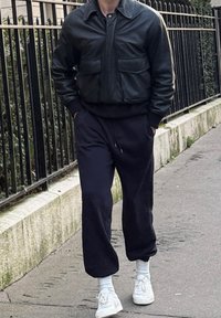 Άτομο που περπατάει στο πεζοδρόμιο φορόντας μαύρο δερμάτινο μπουφάν, μαύρο παντελόνι, άσπρες κάλτσες και άσπρα αθλητικά παπούτσια δίπλα σε μαύρο μεταλλικό φράχτη και πέτρινο τοίχο.