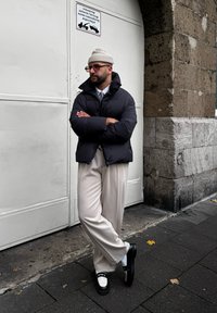 Μαύρο μπουφάν με ψηλό γιακά, μπεζ πλαϊνές παντελόνες και μαύρα παπούτσια με λευκές λεπτομέρειες. Ουδέτερο φόντο από πέτρα και λευκή πόρτα.