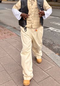 Beige rutig skjorta med knappar, svart läderväst, krämfärgade vida byxor och bruna träskor; har en berlock fäst vid byxorna.