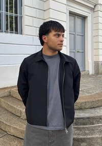 Čierna zipsová bunda cez šedé tričko, so hladkou textúrou a priliehavým strihom. Pozadie zahŕňa kamenné schody a svetlošedé dvere.