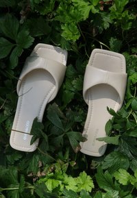 Bežna drsna sandala s kvadratnim nosom in širokim trakom. Gladek sintetičen material z detajli šivanja, ki leži na zeleni vegetaciji.