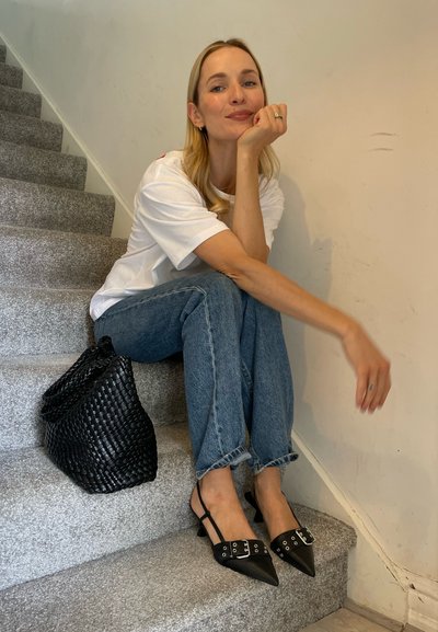 Zapatos slingback negros con punta afilada, adornados con hebillas plateadas, combinados con jeans azules y un bolso negro de rafia, sobre escaleras grises.