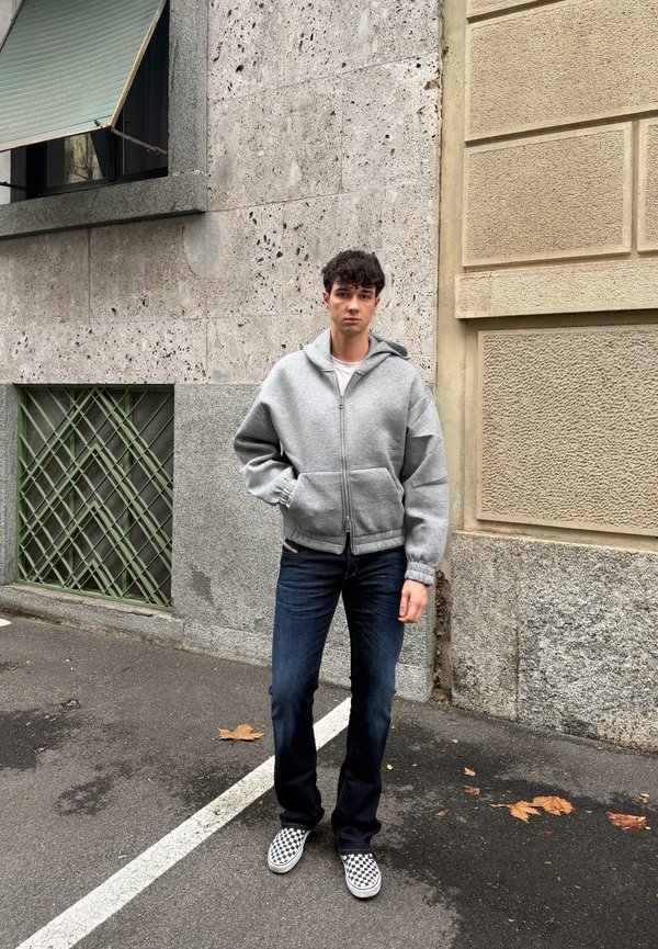 Mladý muž v sivej mikine na zips, tmavých džínsoch a károvaných slip-on topánkach stojaci na mestskom chodníku pri textúrovaných kamenných stenách.