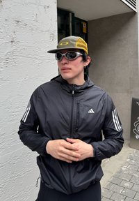 Jeune personne portant une veste Adidas noire, une casquette grise et jaune, et des lunettes de soleil sportives blanches, debout près d'un mur blanc texturé en extérieur.