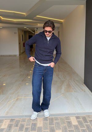 Suéter de punto azul marino con cremallera, camiseta interior gris claro, pantalones de mezclilla azul y zapatillas grises; de pie en un pasillo bien iluminado con suelo de mármol.