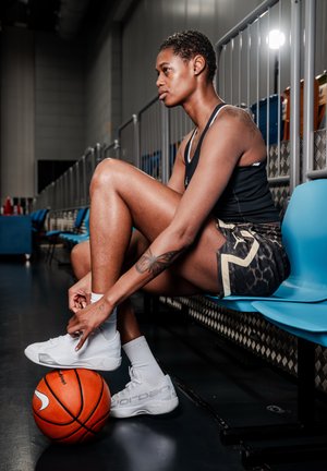 Atleta sentado en las gradas del gimnasio atándose las zapatillas blancas, con una pelota de baloncesto bajo el pie, vistiendo una camiseta sin mangas negra y pantalones cortos con estampado.