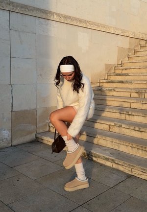 Mujer con suéter blanco peludo y cinta en la cabeza sentada en unas escaleras al aire libre, ajustándose unos zapatos beige sin cordones y calcetines blancos, con un pequeño bolso marrón.