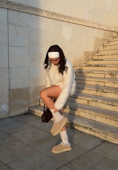 Mujer con suéter blanco peludo y cinta en la cabeza sentada en unas escaleras al aire libre, ajustándose unos zapatos beige sin cordones y calcetines blancos, con un pequeño bolso marrón.