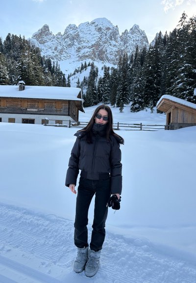 Mujer con chaqueta negra, pantalones y gafas de sol, de pie en un camino nevado con cabañas de madera, altos pinos y montañas nevadas al fondo.