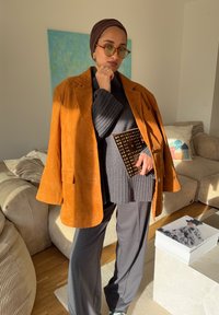 Ohlapen rjavi blazer iz semiša, sivi rebrasti pulover, sive široke hlače, zlato poudarjena torbica, okrogla zelena sončna očala, sproščeno okolje.