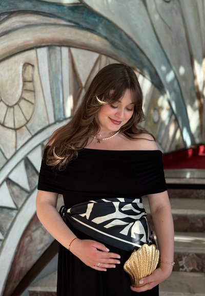 Vestido negro sin hombros con detalles fruncidos, combinado con un fajín de patrón en blanco y negro y un clutch dorado en forma de concha.