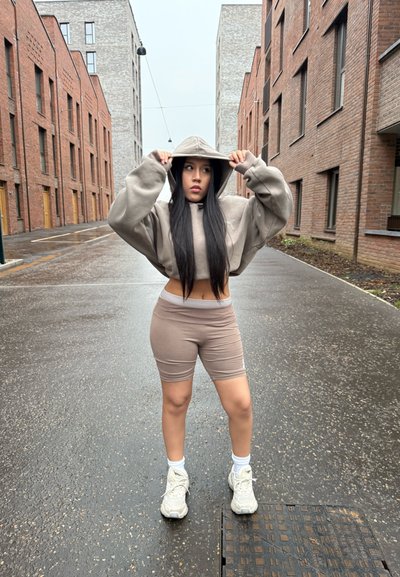 Sudadera corta gris claro y pantalones cortos beige a juego, combinados con zapatillas blancas, de pie en una calle mojada flanqueada por edificios modernos de ladrillo.