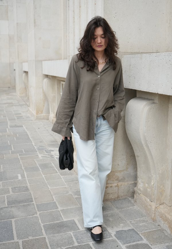Woman with long dark hair wearing a loose olive shirt and light jeans, walking on a stone pavement beside a beige stone wall, holding a black bag.
