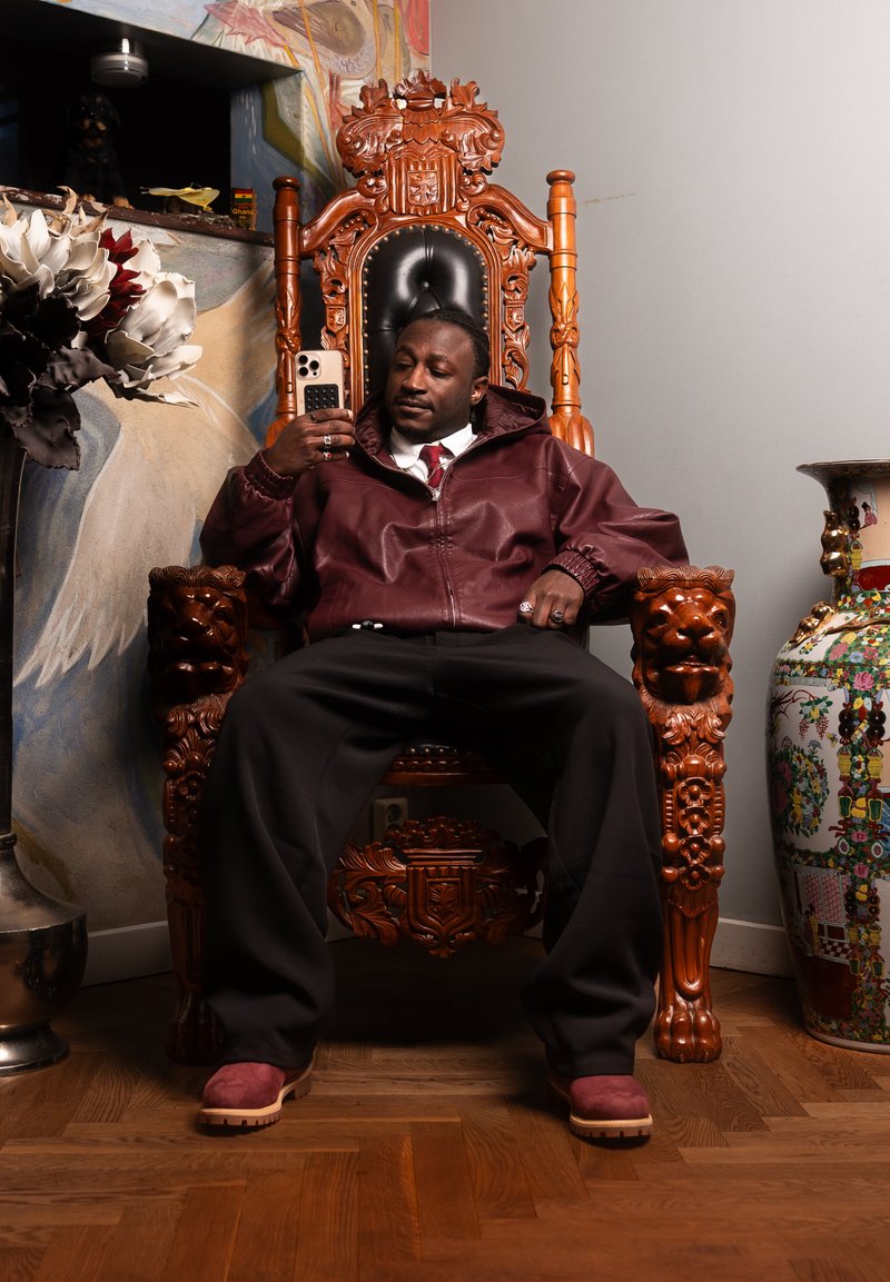 Muž v bordovej bunde sediaci na ozdobnej drevenej trónovej stoličke, drží smartfón, za ním veľká kvetinová váza a dekoratívna stena.