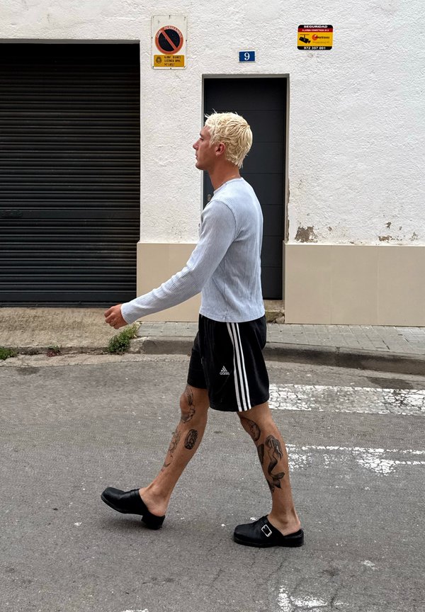 Maglietta a maniche lunghe ribbed blu chiaro, pantaloni corti neri con strisce bianche, zoccoli neri e tatuaggi visibili sulle gambe, che cammina per strada.