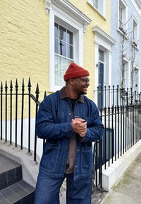 Geacă și pantaloni din denim albastre închise, cu un guler din catifea maro, asortați cu o căciulă roșie. Ochelari negri și un pulover texturat pe dedesubt.