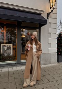 Manteau camel sur un pull blanc en maille torsadée, pantalon beige à jambes larges et bottes à plateforme camel. Tenant une tasse de café festive.