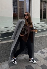 Cappotto di lana grigio sopra un maglione marrone a coste, leggings neri e sneakers bianco e nere. Tiene in mano una tazza di caffè di carta marrone e una borsa marrone.