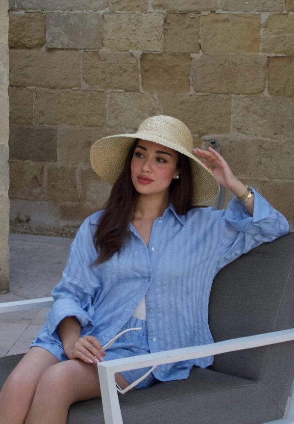 Woman in a blue outfit and wide-brimmed straw hat sits on a gray chair against a stone wall, holding sunglasses in one hand.