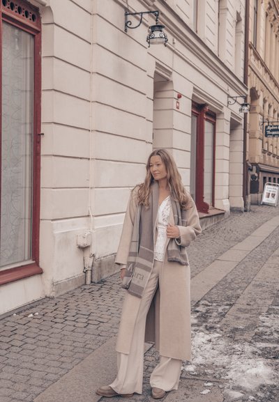 Mujer con abrigo beige, bufanda y conjunto blanco, de pie en una calle empedrada cerca de edificios históricos con ventanas con bordes rojos.