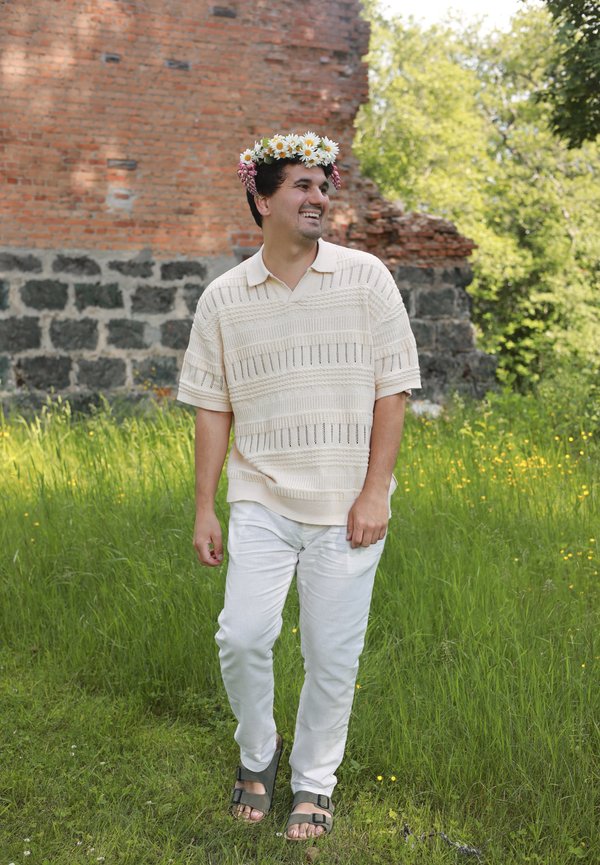 Mann mit Blumenkrone und heller Kleidung, der lächelnd auf Gras in der Nähe einer Ziegel- und Steinmauer im Freien geht.