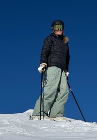 Conjunto de esqui com um casaco puffer preto e calças de neve verde claro. Óculos de esqui pretos com lentes refletivas, segurando bastões de esqui pretos na neve.
