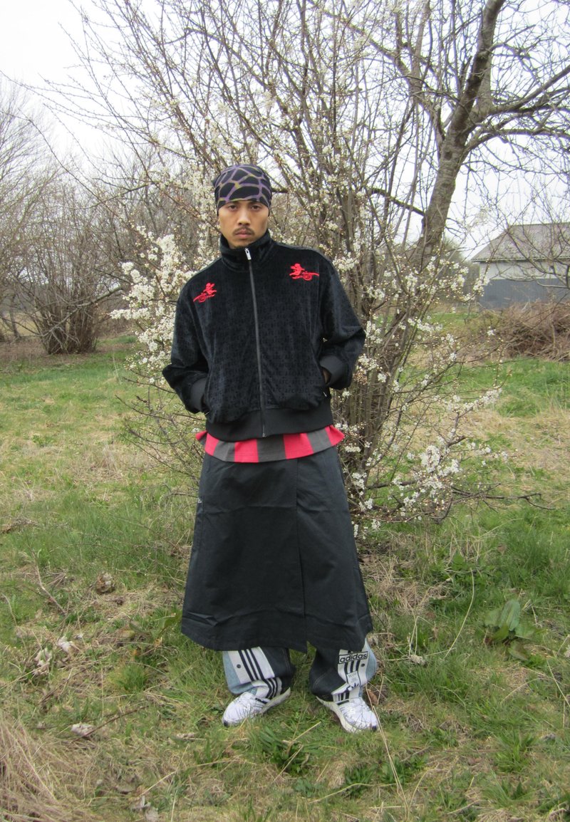 Un homme se tient à l'extérieur, vêtu de vêtements superposés noirs et rouges, d'un couvre-chef à motifs et de baskets blanches Adidas, avec des arbres sans feuilles et des branches en fleurs derrière lui.