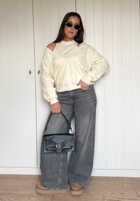 Kremast pulover z odkrito ramo, sive široke jeans hlače, bež platformni škornji, drži črno usnjeno torbico z zlatim zaponko.