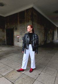 Must mustaari nahast mootorratturi jope, sügavalt sümmeetriline must särk, valged laiad püksid ja punased võrgust kingad rihmadega. Seisab plaaditud kõnniteel.