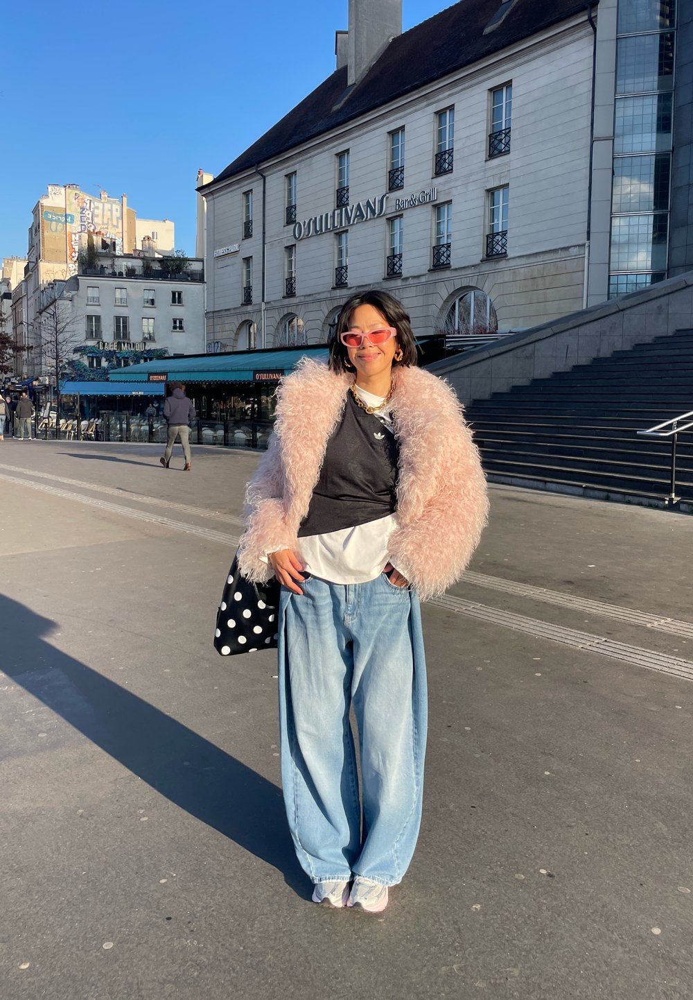 Personne portant une veste rose duveteuse, un jean bleu oversize, des lunettes de soleil roses et tenant un sac à pois, debout à l'extérieur près du Bar & Grill O'Sullivan.