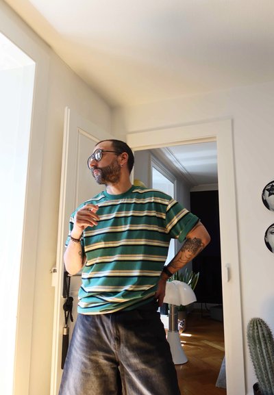 Camisa de manga corta a rayas en tonos verde azulado y crema, combinada con pantalones cortos de mezclilla oscuros. El fondo presenta plantas de interior y una lámpara.